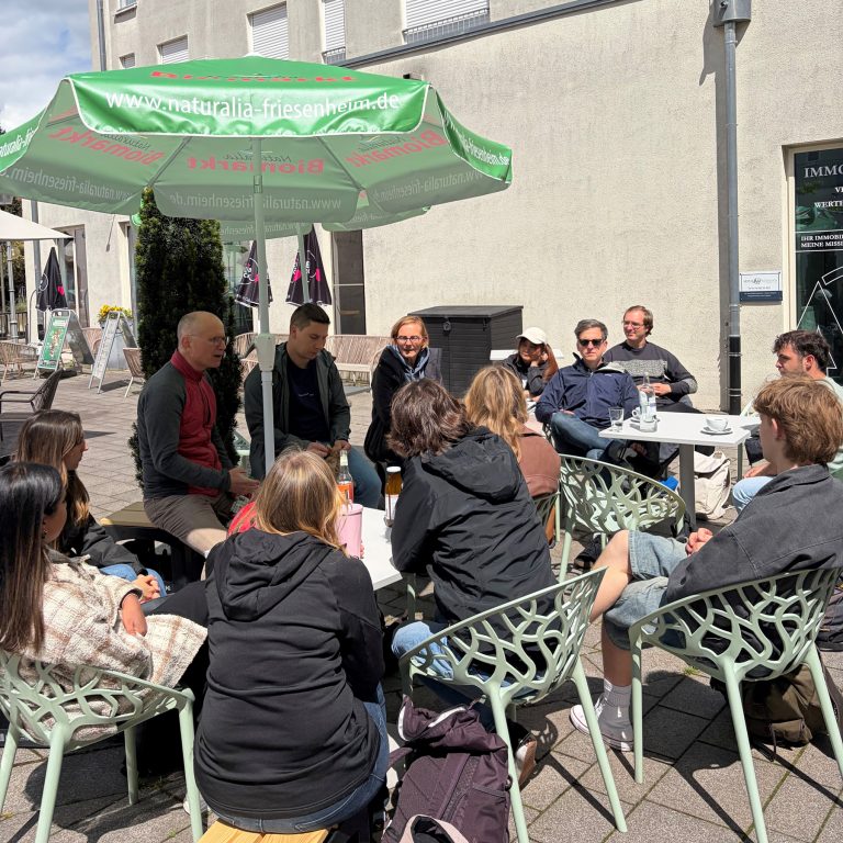 Studierende aus Freiburg sitzen mit Irene Krieg vom Naturalia und ihren Professoren unter einem Sonnenschirm im Freien an Tischen im Gespräch